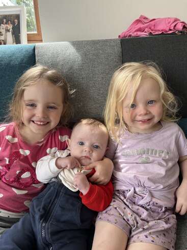 Three young children sit closely on a sofa, smiling warmly. The two older girls wear colorful shirts, holding a baby between them. A family photo decorates behind.