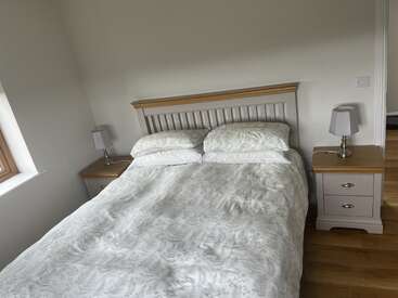 This image shows a neatly made bed with a wooden headboard, two matching bedside tables with lamps, light bedding, and wooden flooring in a bright room.