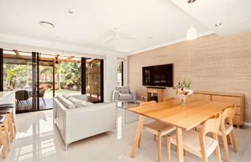 Cette image montre un salon et une salle à manger modernes et lumineux, avec des meubles en bois clair, un salon blanc, des plantes d'intérieur, de grandes portes coulissantes en verre et une vue sur un jardin.