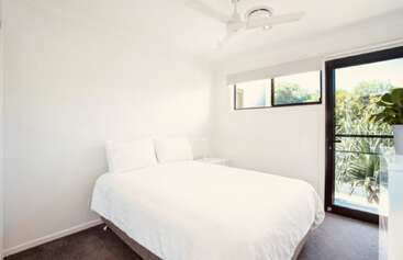 Il s'agit d'une chambre à coucher lumineuse et minimaliste avec des murs blancs, un lit double, un ventilateur de plafond, une fenêtre, une porte vitrée, une table de chevet et un peu de verdure à l'extérieur. Ambiance paisible.