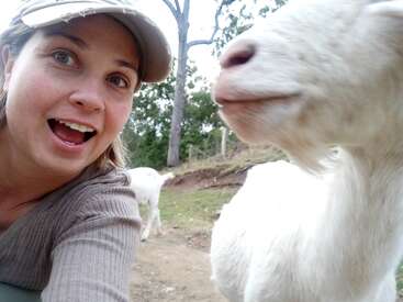 La imagen muestra a una mujer con gorra y top gris, tomándose un selfie con una cabra blanca en un campo, con otra cabra al fondo.