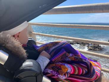 Un bébé, chaudement emmitouflé dans un tissu coloré, est assis dans une poussette sur une promenade, regardant les vagues bleues et ensoleillées de l'océan à travers les grilles.