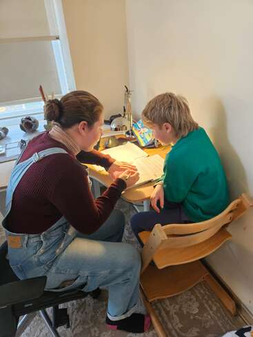 Une femme aide un enfant à faire ses devoirs à un bureau. Ils sont tous deux concentrés, utilisant des crayons et du papier, entourés de livres et d'une lampe lumineuse.