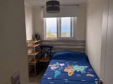 Une petite chambre confortable avec un lit simple recouvert d'une couverture de carte du monde, des étagères en bois, une chaise, une grande fenêtre et un plafonnier moderne.
