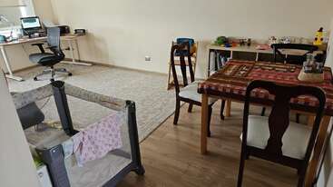 Cette image montre une pièce multifonctionnelle avec un espace de travail, une table à manger, un parc pour enfants, des chaises, des étagères et divers objets, mélangeant les espaces de bureau, de vie et de jeu pour les enfants.