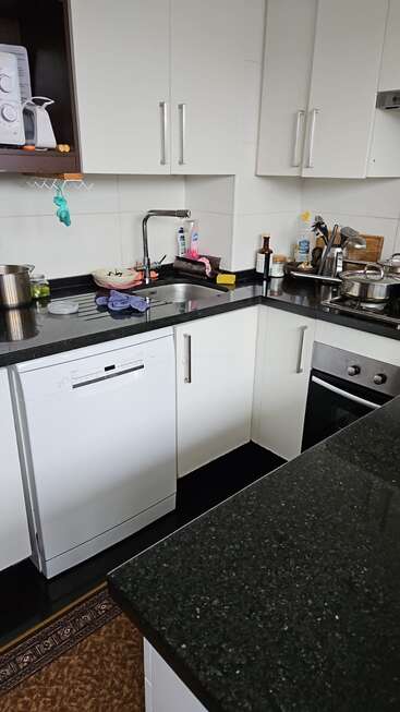 Il s'agit d'une cuisine moderne avec des armoires blanches, un lave-vaisselle, un four, un plan de travail encombré de vaisselle, d'ustensiles et de produits de nettoyage, et plusieurs casseroles à côté de l'évier.