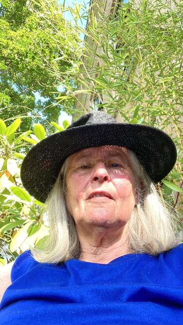 A woman with long gray hair wears a black wide-brimmed hat and blue shirt, sitting outside surrounded by green plants and sunlight, enjoying a sunny day.