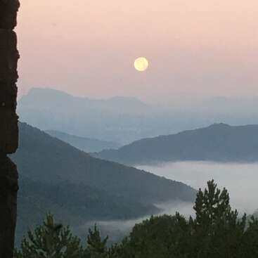 Une pleine lune plane sur des montagnes brumeuses au crépuscule, jetant une lueur sereine. Des pins au premier plan, avec des dégradés de rose et de bleu dans le ciel.