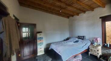 La imagen muestra una habitación con una cama, una cómoda y una mesilla de noche, con un edredón blanco y almohadas, y una ventana con cortinas al fondo. La habitación está bien iluminada y organizada.