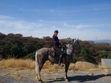 Una mujer está sentada sobre un caballo gris al aire libre, rodeada de árboles, rocas y hierba seca. Sonríe y hace el gesto de la paz bajo un cielo despejado.