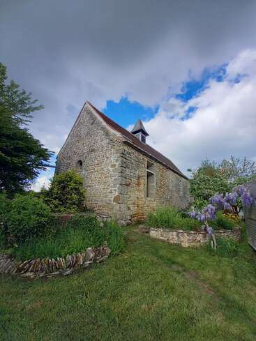 La imagen representa una iglesia de piedra con tejado y campanario marrones, rodeada de exuberante vegetación y vibrantes flores moradas en un día nublado.