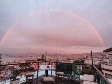Un impresionante arco iris cruza un cielo rosado y nublado sobre un paisaje urbano. En primer plano, una acogedora azotea con sillas y una mesa.
