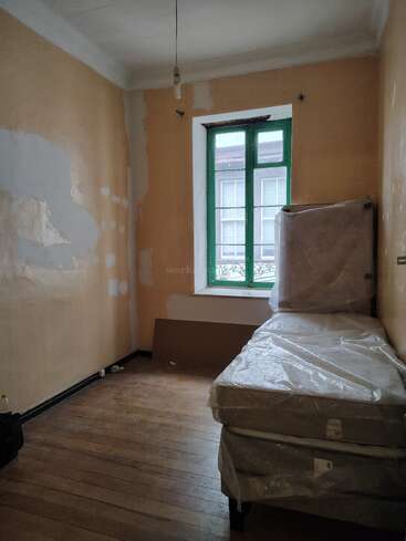 Esta imagen muestra una habitación pequeña y escasamente amueblada, con paredes sin terminar, una sola bombilla, dos colchones envueltos en plástico, suelo de madera y una ventana con un ribete verde.