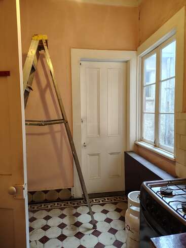 En un estrecho rincón de la cocina hay una puerta blanca cerrada, una escalera metálica, suelo de baldosas a cuadros, grandes ventanas, una cocina de gas, una bombona de propano y paredes de color melocotón.
