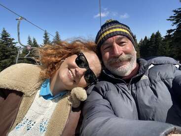 Un couple souriant prend un selfie en plein air par une journée ensoleillée. Ils portent des vestes et des bonnets d'hiver, avec des arbres et un ciel bleu en arrière-plan.