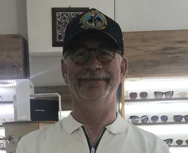 Un homme portant des lunettes et une casquette bleu marine se tient dans un magasin de lunettes, entouré d'étagères de lunettes de soleil, souriant légèrement, vêtu d'une chemise blanche à col.