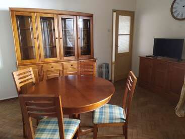 The image shows a dining room with a round wooden table, four chairs with blue plaid cushions, a glass cabinet, TV, clock, and wooden sideboard.