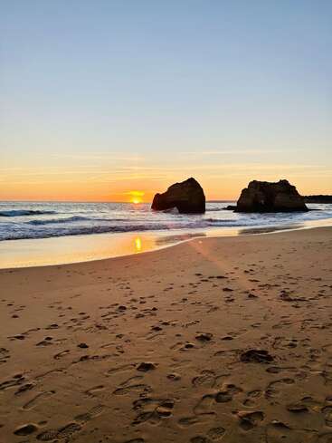 Golden sunset over a tranquil beach, with gentle waves, wet sand reflecting sunlight, large rock formations in the water, and scattered footprints across the peaceful shoreline.