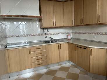 This is a clean, empty kitchen with wooden cabinets, tiled walls, a stainless steel sink, marble countertops, and beige checkered flooring. There is also a shelf.