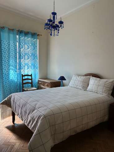 This bedroom features a neatly made bed with checkered bedding, blue patterned curtains, a wooden chair, nightstand, blue lamp, and a striking blue chandelier.