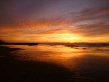 Ein atemberaubender Sonnenuntergang beleuchtet den ruhigen Strand und taucht den Himmel und den nassen Sand in leuchtende Orange-, Gold- und Violetttöne, die eine ruhige Atmosphäre schaffen.