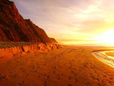 Goldenes Sonnenlicht durchflutet einen Sandstrand und hinterlässt Fußabdrücke entlang des Ufers. Auf der linken Seite erheben sich steile Klippen, während sanfte Wellen den warmen, glühenden Sonnenuntergang reflektieren.