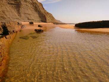 Klares, seichtes Wasser fließt zwischen felsigen Klippen und sandigen Ufern unter einem hellen Himmel. Das Sonnenlicht glitzert auf dem Wasser und schafft eine friedliche, ruhige Strandszene.