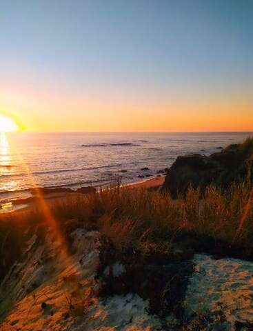 Das Bild zeigt einen wunderschönen Sonnenuntergang über dem Meer, der ein goldenes Licht auf die ruhigen Wellen, sandigen Klippen und grasbewachsenen Dünen wirft und so eine friedliche, ruhige Szene schafft.