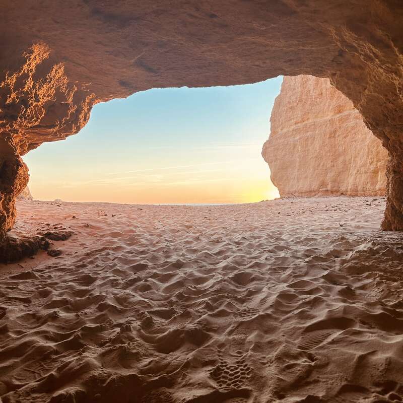 Viewed from inside a sandy cave, the image shows a bright, serene beach and calm sky at sunset, with warm sunlight illuminating the cave walls.