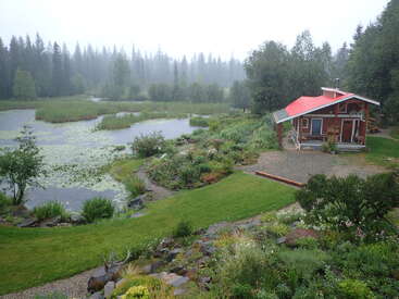 A cozy wooden cabin with a red roof sits beside a tranquil pond, surrounded by lush greenery, gardens, and towering trees on a misty day.