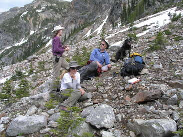 Drei Menschen und ein schwarzer Hund ruhen sich an einem felsigen Berghang aus. Sie tragen Wanderkleidung, sind von Rucksäcken, kleinen Bäumen und Schneeflecken umgeben und genießen die Natur.