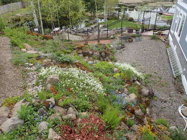 Ein abfallender Steingarten mit blühenden Blumen und viel Grün, neben einem Kiesweg und einer Terrasse, bietet einen Blick auf einen umzäunten Garten in der Nähe eines Teiches mit einem Steg.