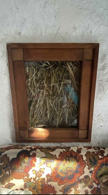 A imagem mostra uma parte de uma parede com um painel de vidro com moldura de madeira que mostra palha por dentro. Abaixo dele, tem uma almofada estofada colorida e estampada.