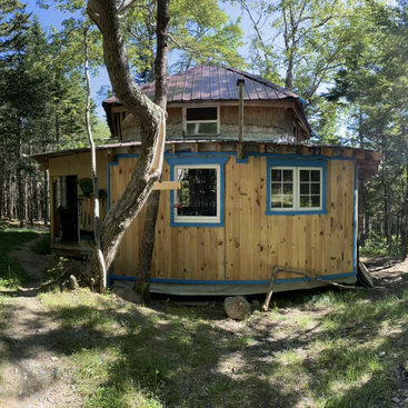 Esta imagen muestra una pequeña cabaña redonda de madera con techo metálico, ventanas con marcos azules y rodeada de árboles en un tranquilo entorno forestal.