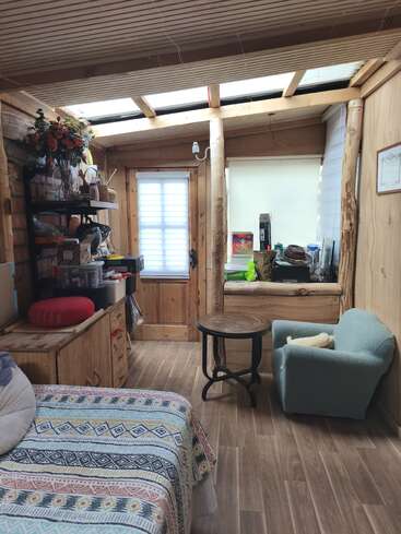 This cozy wooden room has a patterned bedspread, armchair, round table, shelves, flowers, and skylights, creating a warm, rustic, and inviting atmosphere.