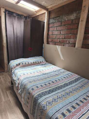 This cozy room features a single bed with a colorful patterned blanket, wooden beams, exposed brick wall, wardrobe, curtain, skylight, and light wood flooring.