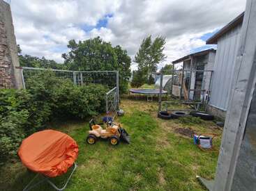 A grassy backyard with a toy excavator, orange chair, trampoline, soccer ball, and tires. Surrounded by bushes, metal fencing, and buildings under a cloudy sky.