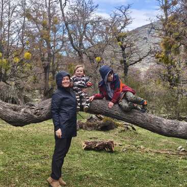 A woman and two children enjoy an outdoor adventure, sitting and standing on a horizontal tree trunk surrounded by leafless trees and mountains under a cloudy sky.