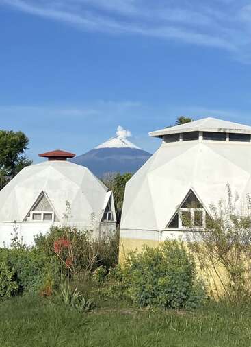 Duas casas de domo geodésico branco estão entre a folhagem verde. Ao fundo, um vulcão coberto de neve emite fumaça contra um céu azul claro, criando uma cena pacífica.