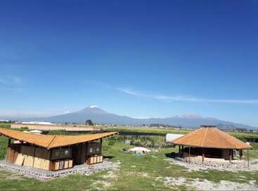 Deux bâtiments rustiques en bois aux toits de tuiles rouges se dressent dans un champ verdoyant, avec en arrière-plan des montagnes et un volcan enneigé qui s'élèvent majestueusement sous un ciel bleu limpide.