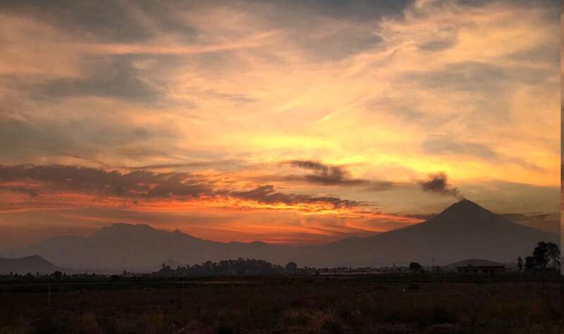 La imagen representa un paisaje sereno con una majestuosa cadena montañosa, con un pico prominente en el centro, rodeado de exuberante vegetación y un cielo azul despejado. La escena irradia tranquilidad y belleza natural.