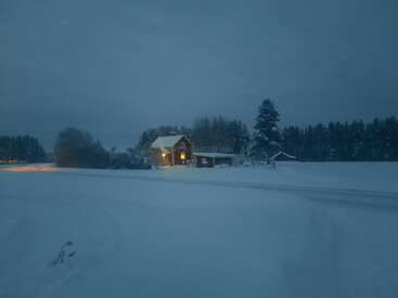A cozy house glows warmly in a snowy, serene landscape surrounded by trees under a gray sky, evoking a peaceful winter evening in the countryside.