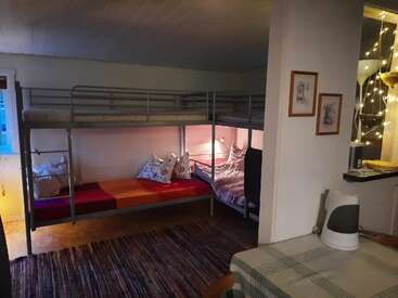 A cozy room with a bunk bed, colorful bedding, pillows, rug, table with a kettle, framed pictures, string lights, and a warm, homely atmosphere.