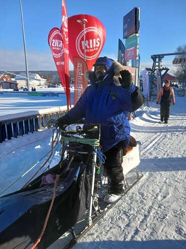 A person dressed in warm winter clothing stands on a sled, smiling and waving. Snow covers the ground, with colorful flags and banners in the background.