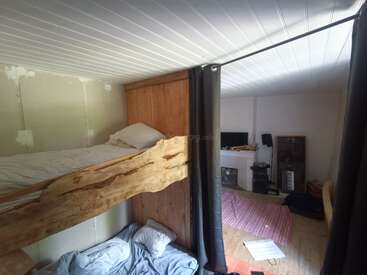 Esse quarto aconchegante tem beliches de madeira rústica, uma parede parcialmente acabada, roupas de cama simples, uma TV, instrumentos musicais, um tapete vermelho e papéis espalhados pelo chão.