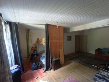 Esta imagem mostra um cômodo espaçoso e um pouco desorganizado com piso de madeira, uma cama pequena, divisória, sofá, tapetes, vigas expostas e luz que atravessa as cortinas.