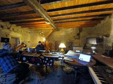 A rustic studio room with wooden beams and stone walls. Two people work among synthesizers, keyboards, laptops, and audio equipment. Soft lighting creates a warm, creative atmosphere.