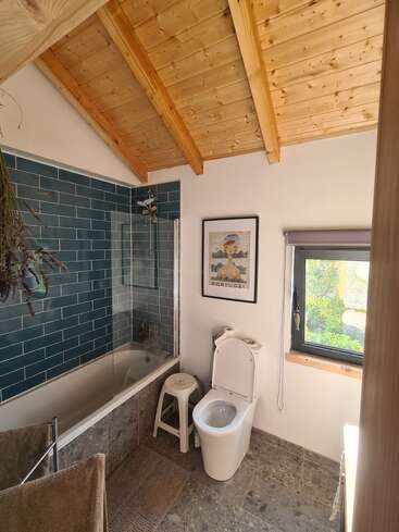 This bathroom features a wooden ceiling, blue tiled bathtub, modern toilet, small window, Portugal poster, white stool, stone floor, and natural light, creating a cozy atmosphere.