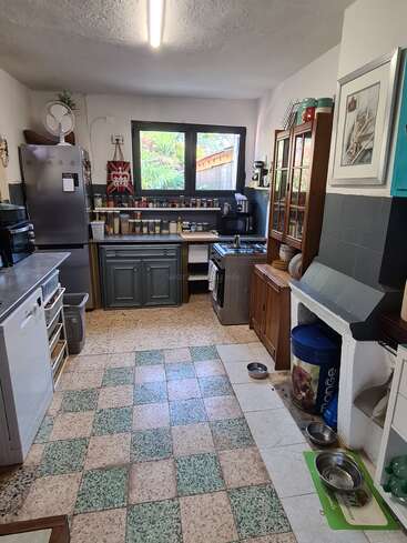 This cozy kitchen features checkered tile flooring, a stainless-steel fridge, wooden cabinets, open shelving, dog bowls, and large windows letting in natural light. It's well-organized and homey.