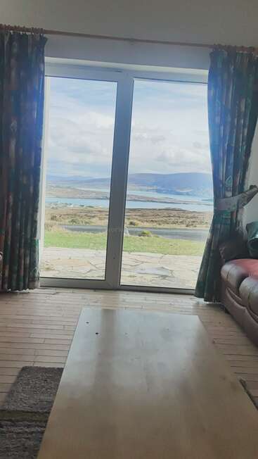 Dieses Bild fängt einen ruhigen Blick durch große Glastüren ein und zeigt Vorhänge, ein Sofa, einen Couchtisch und eine atemberaubende Landschaft mit Hügeln, Himmel und Wasser in der Ferne.
